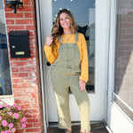 Woman in green overalls and yellow sweater standing on a porch with flowers and a mailbox.