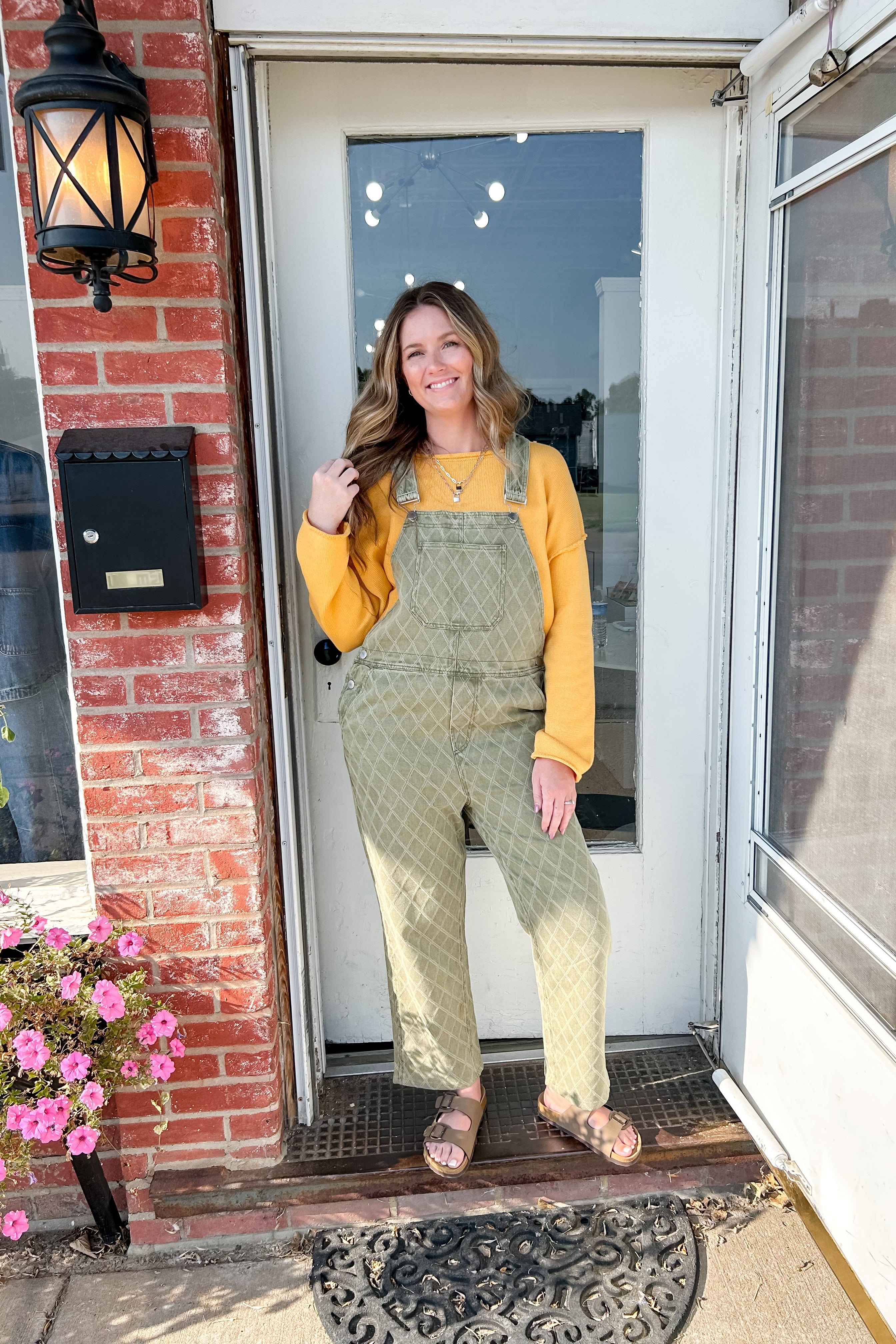 Woman in green overalls and yellow sweater standing on a porch with flowers and a mailbox.