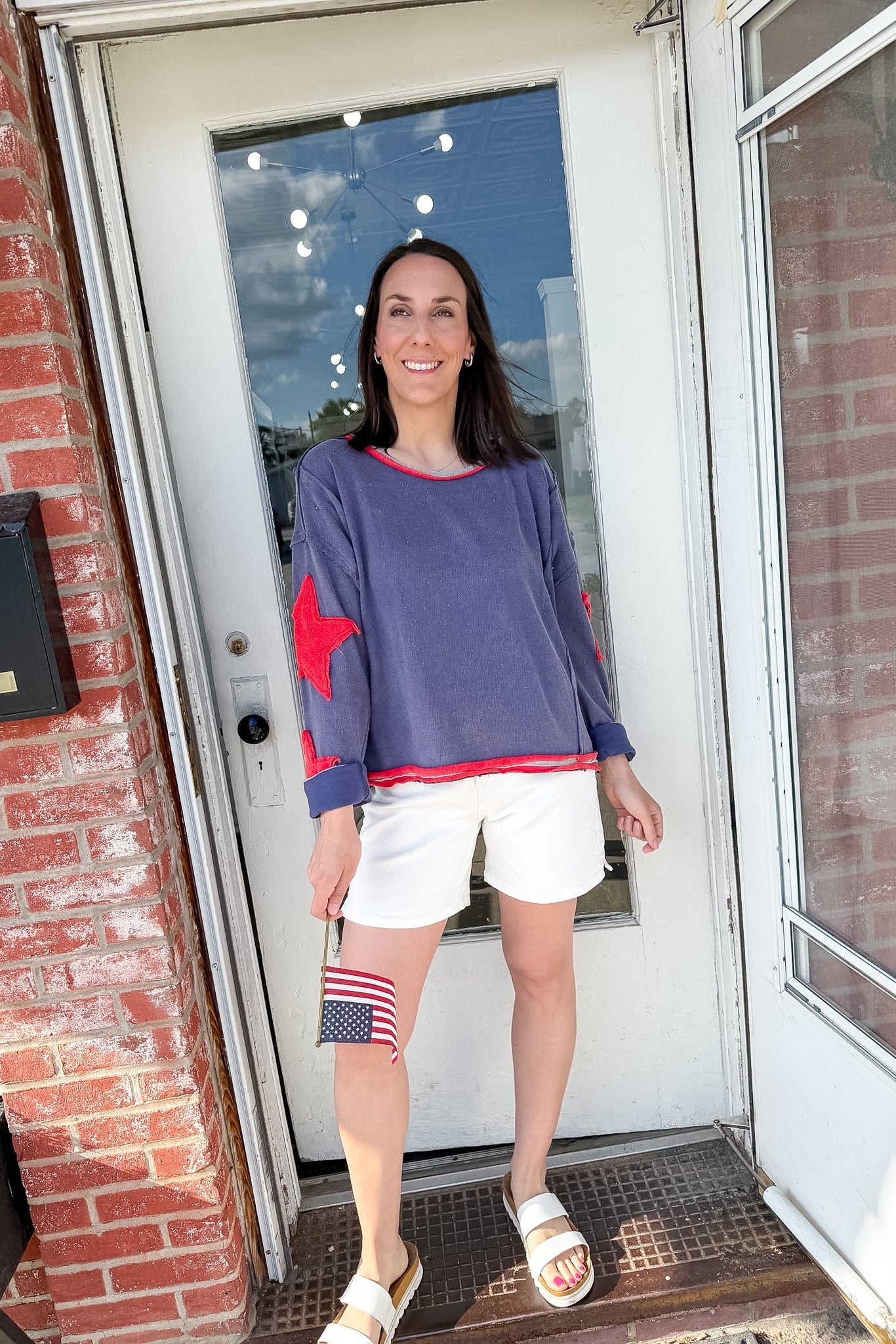 Woman standing outside a house wearing a blue top with red accents and white shorts.