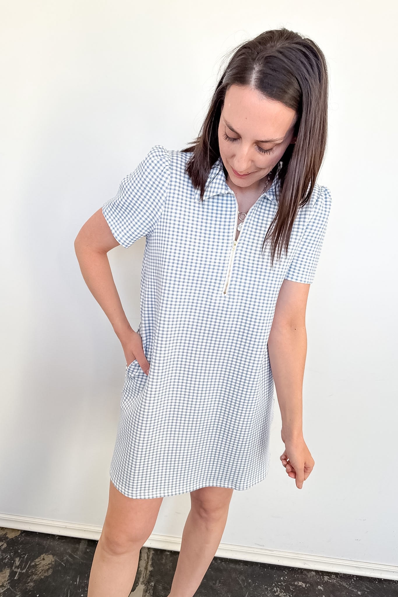 Woman wearing a blue and white checkered dress against a white wall.