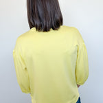 Person wearing a yellow long-sleeve top and blue jeans against a white wall.