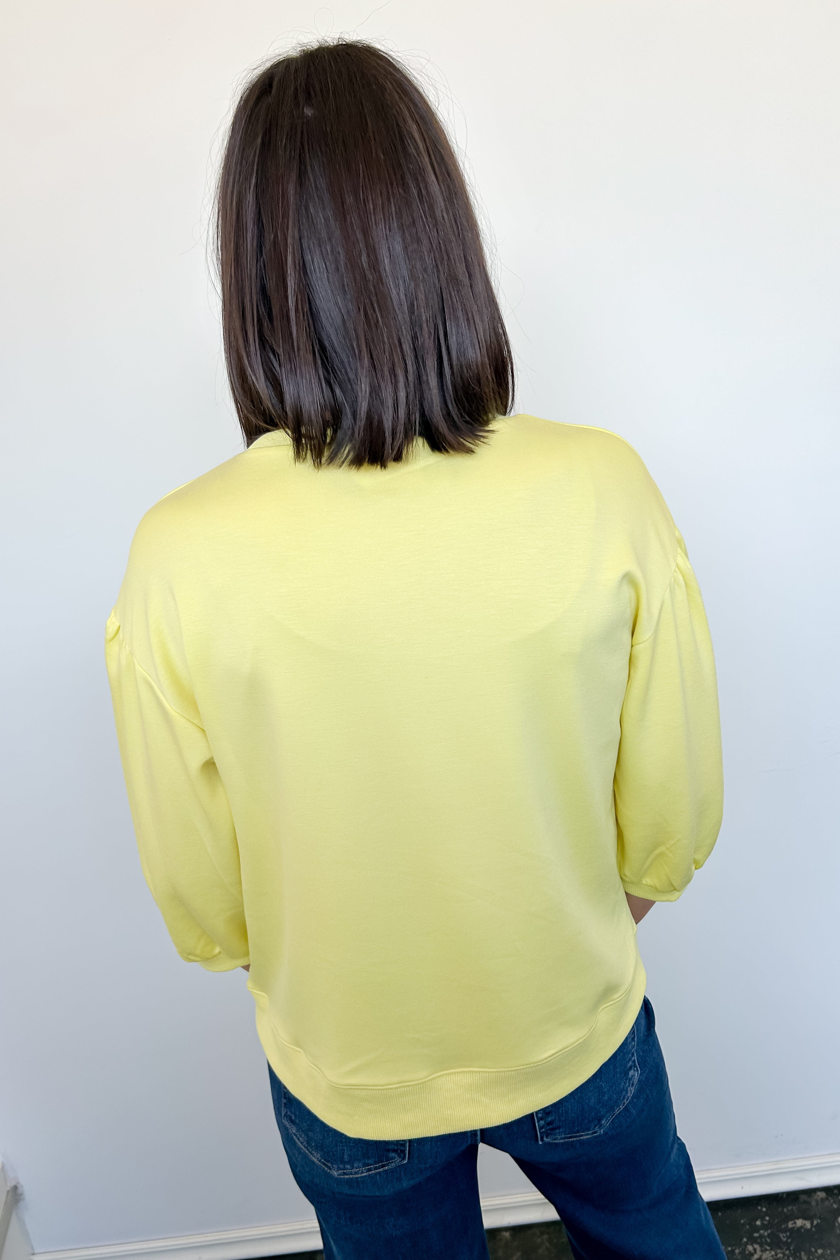 Person wearing a yellow long-sleeve top and blue jeans against a white wall.