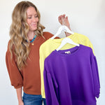 Woman holding three sweatshirts in brown, yellow, and purple against a white wall.