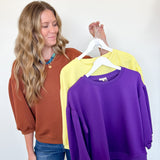 Woman holding three sweatshirts in brown, yellow, and purple against a white wall.