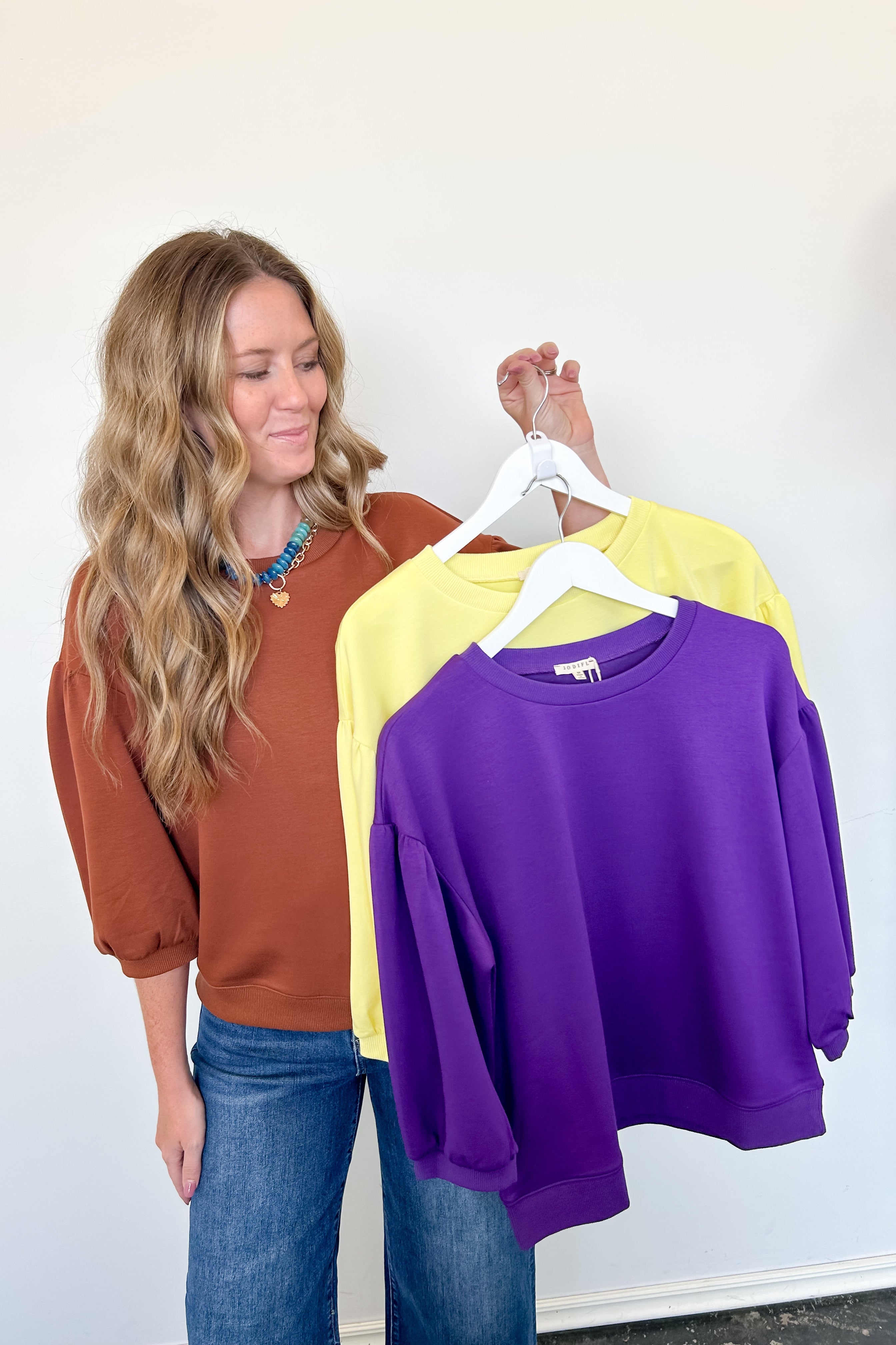 Woman holding three sweatshirts in brown, yellow, and purple against a white wall.