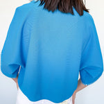 Person wearing a blue textured top with white pants against a plain background