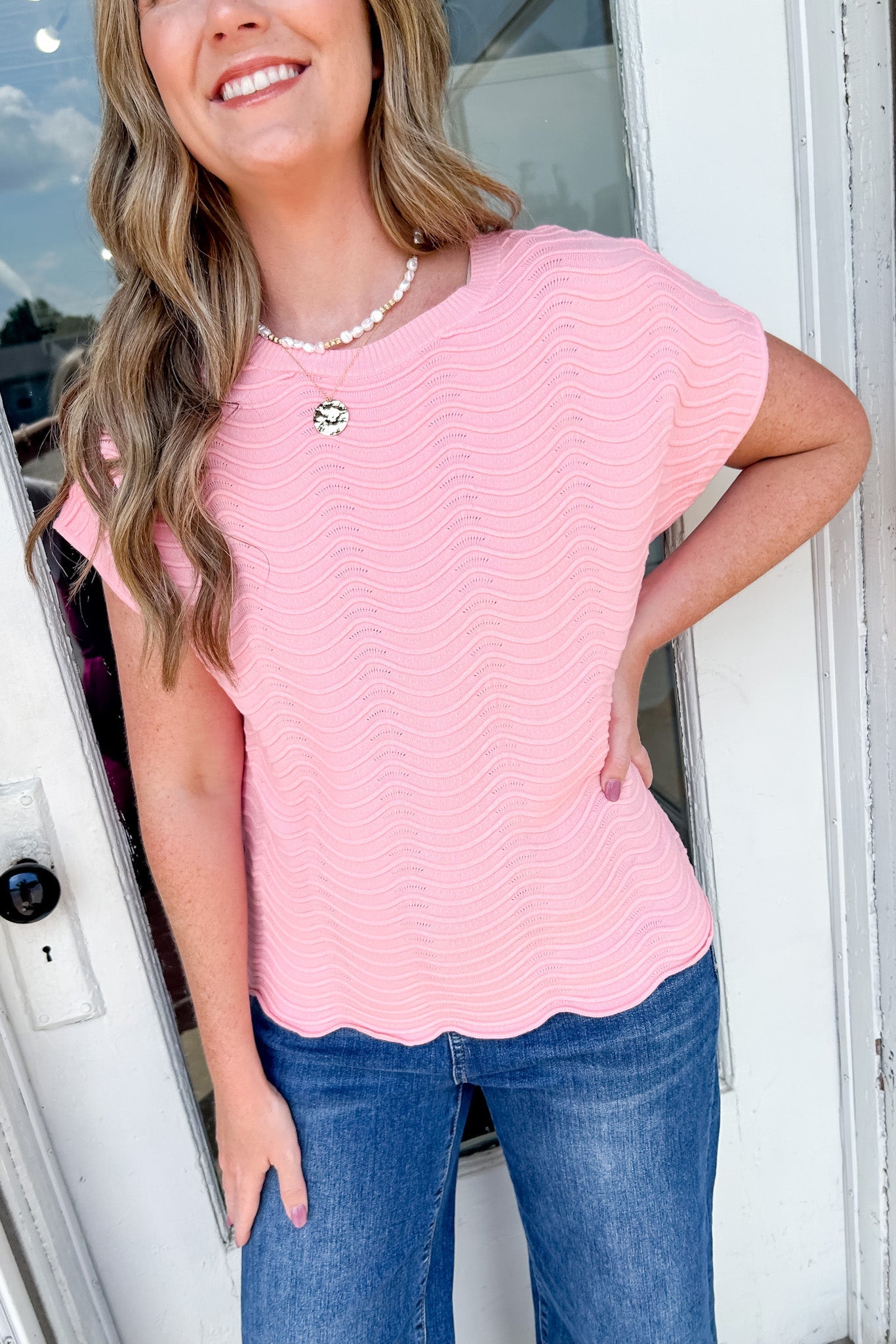 Woman wearing a pink scalloped hem top and blue jeans standing in front of a door.