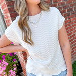 Woman wearing a white textured top and light blue jeans standing in front of a brick wall with flowers.
