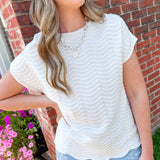Woman wearing a white textured top and light blue jeans standing in front of a brick wall with flowers.