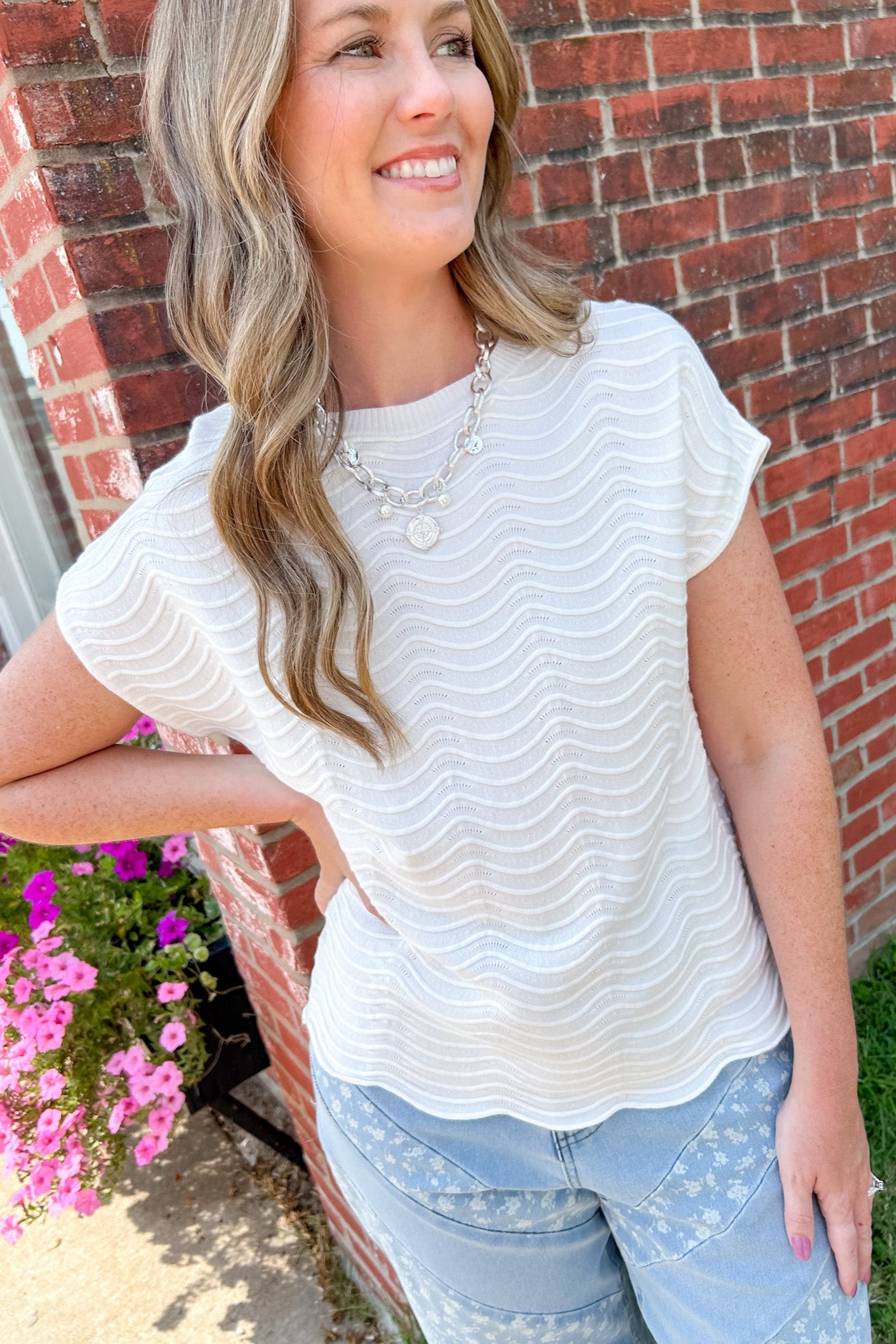 Woman wearing a white textured top and light blue jeans standing in front of a brick wall with flowers.