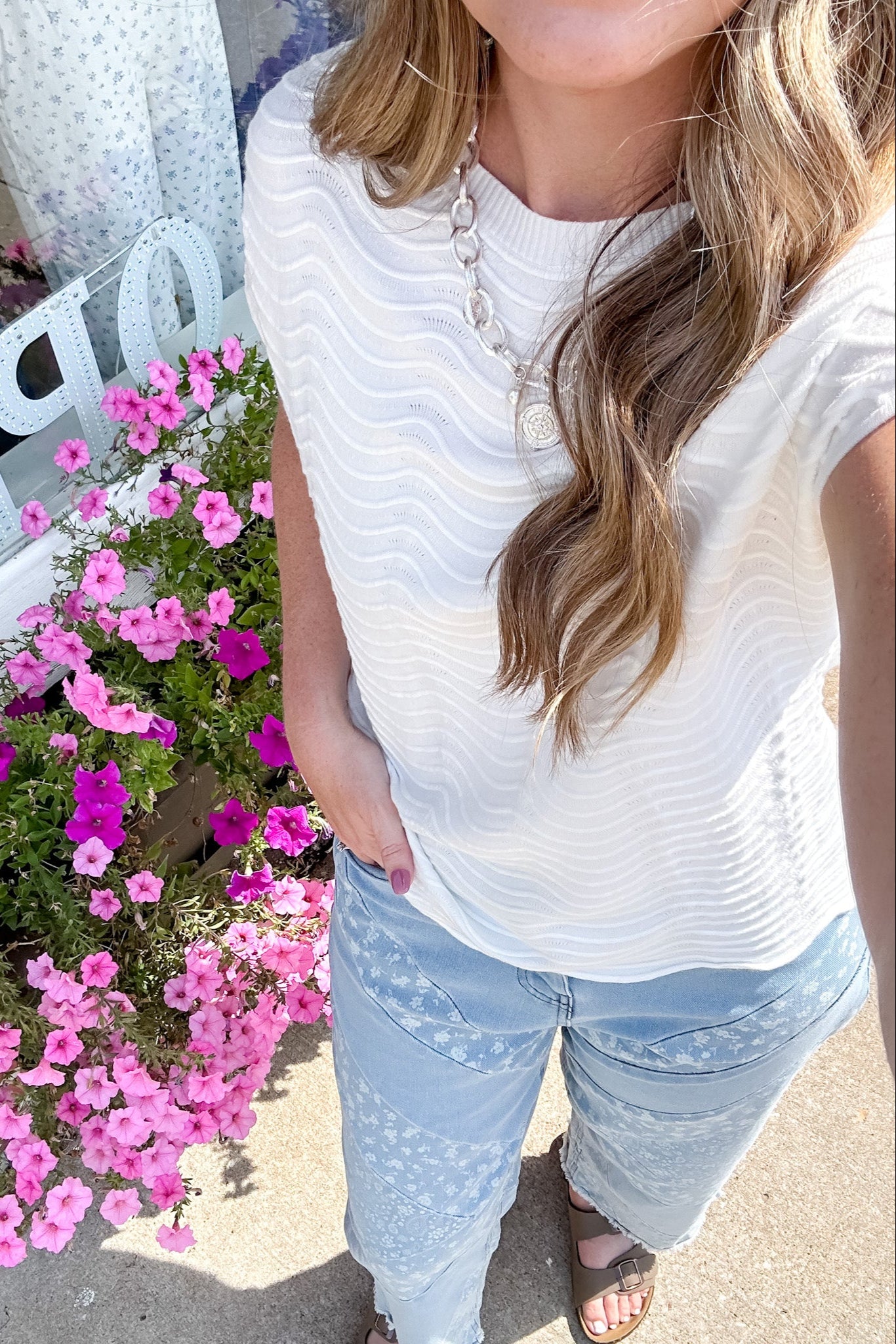 Person wearing a white top and light blue jeans standing next to pink flowers.