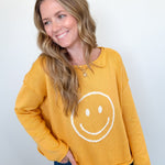 Woman wearing a mustard yellow sweatshirt with a smiley face design against a white wall.