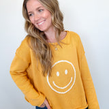 Woman wearing a mustard yellow sweatshirt with a smiley face design against a white wall.