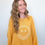Woman wearing a mustard yellow sweatshirt with a smiley face design on a plain background