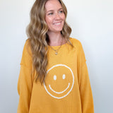 Woman wearing a mustard yellow sweatshirt with a smiley face design on a plain background