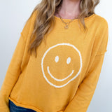 Woman wearing a mustard yellow sweater with a smiley face design against a white background