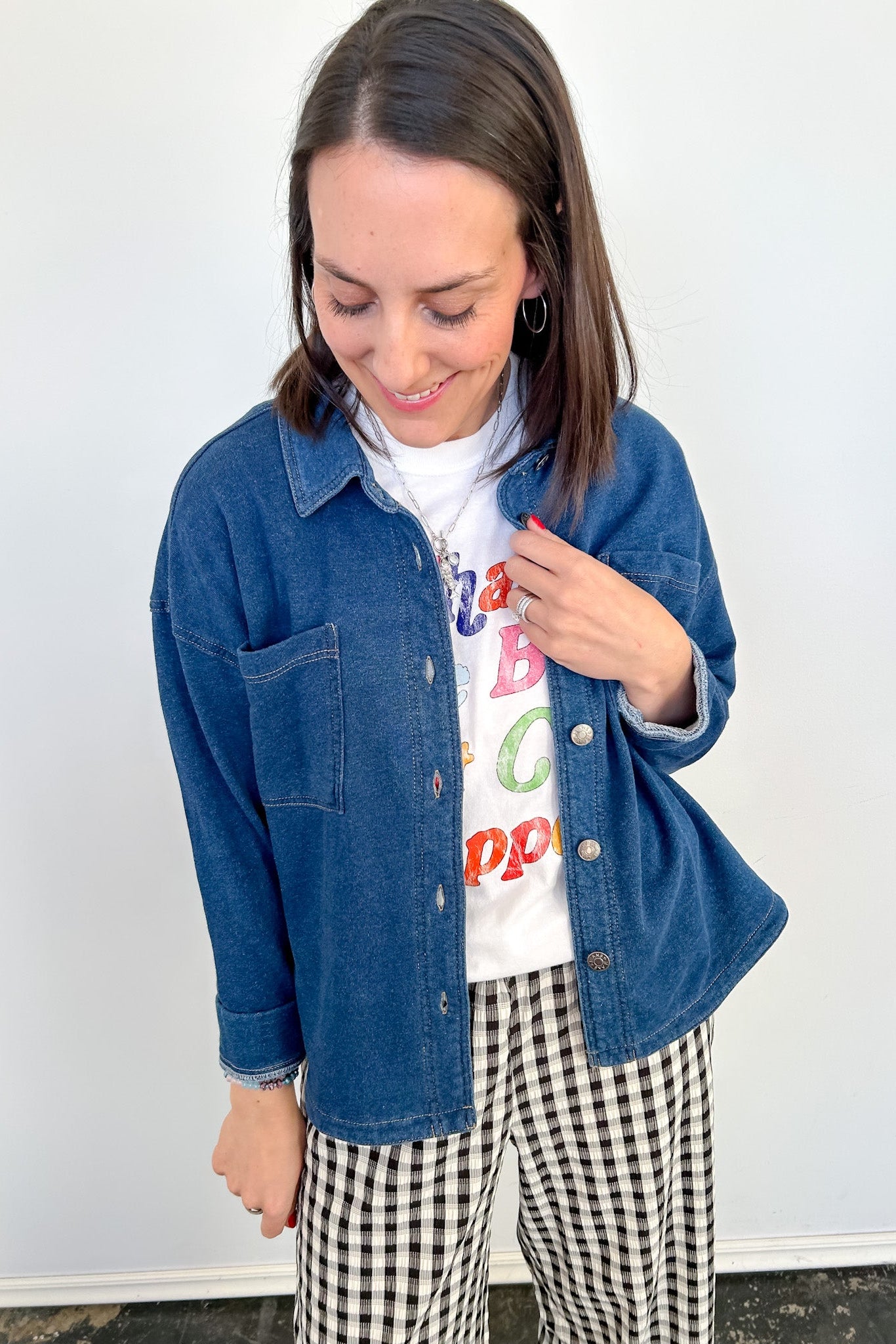 Woman wearing a denim jacket, colorful shirt, and checkered pants against a white wall.