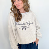 Woman wearing a beige sweatshirt with text and graphics, standing against a white wall.