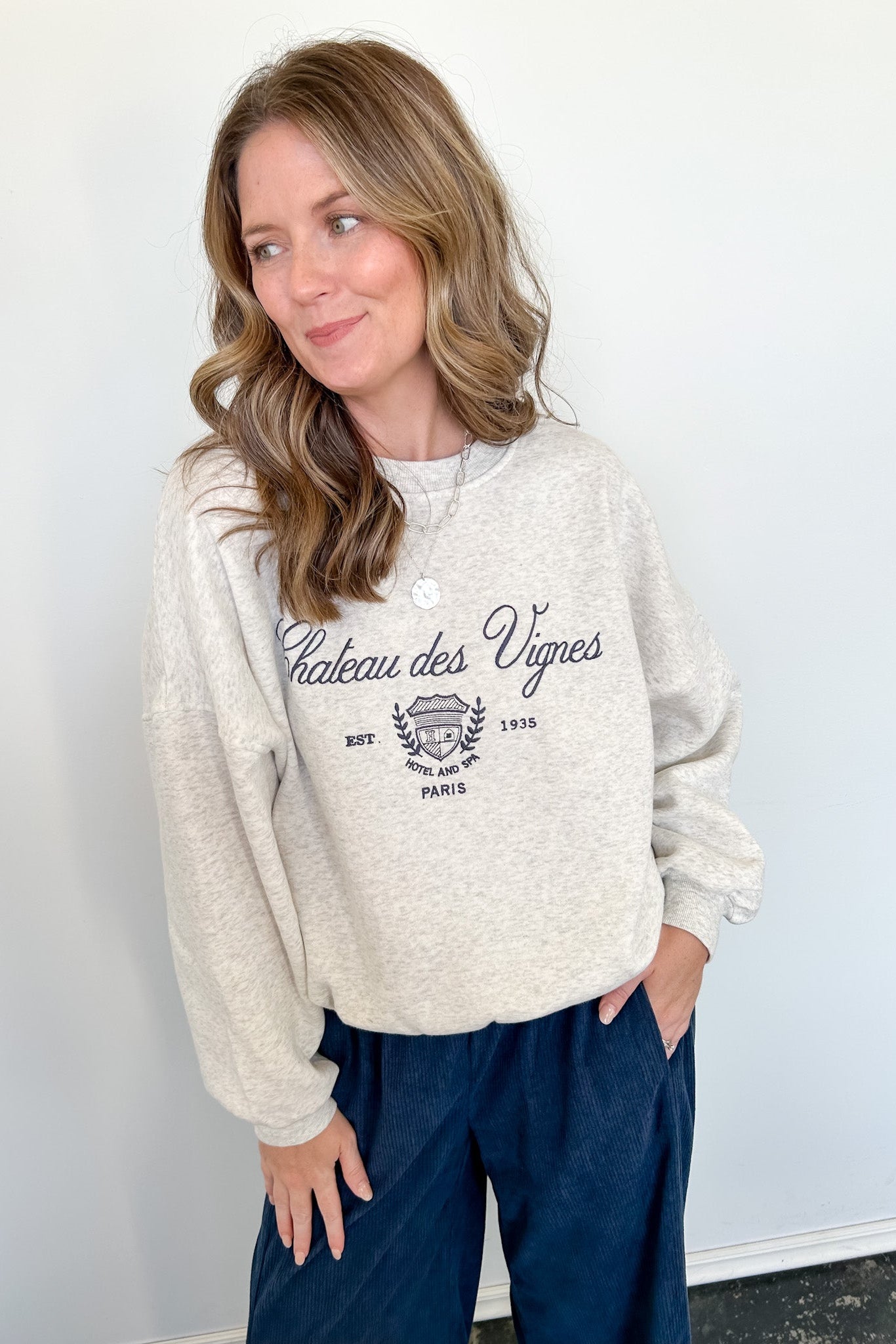 Woman wearing a beige sweatshirt with text and graphics, standing against a white wall.