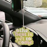Car air freshener with 'the best is yet to come' text hanging from a car's rearview mirror.