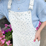 Person wearing a blue sweater and white floral overalls standing outdoors with pink flowers in the background.