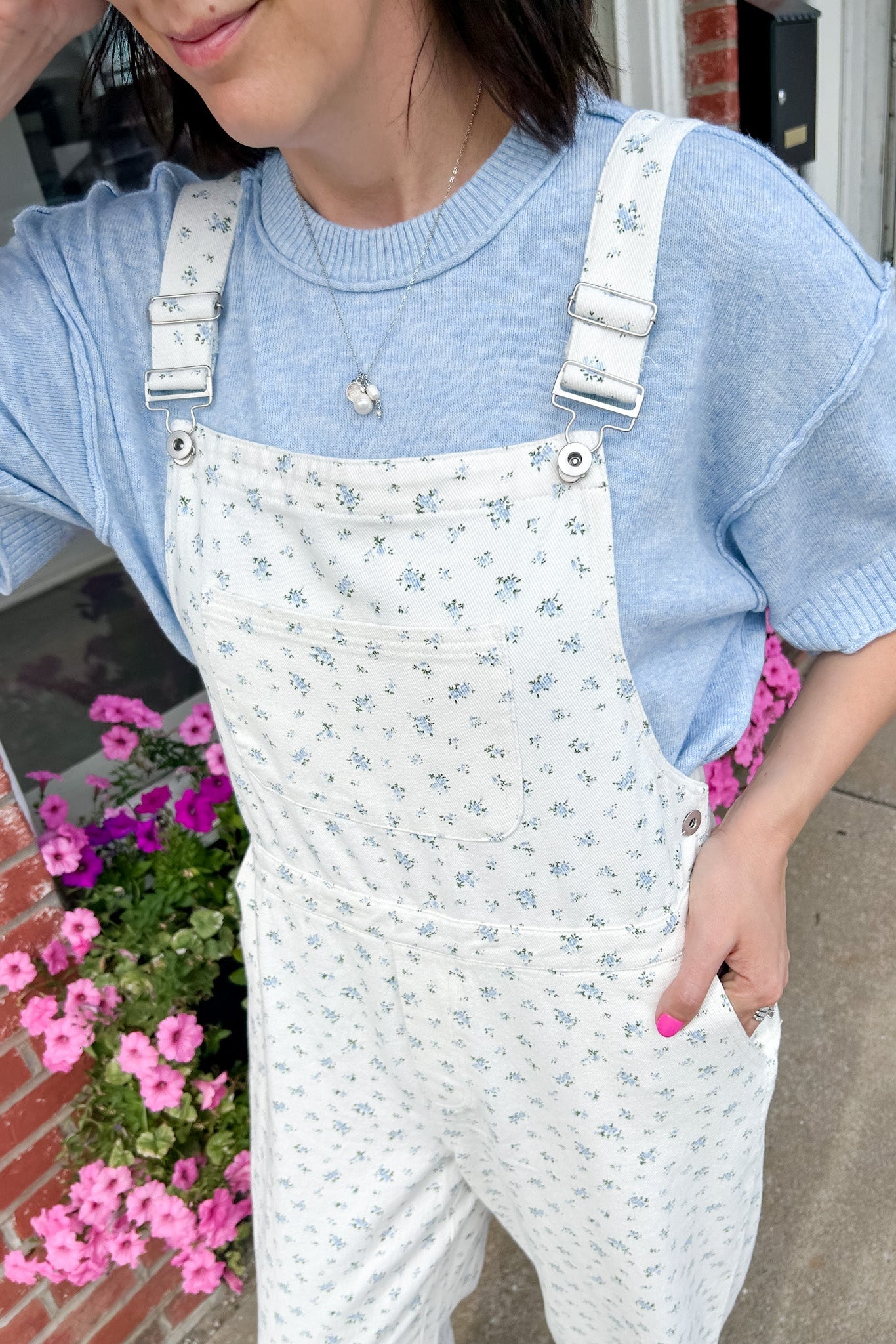 Person wearing a blue sweater and white floral overalls standing outdoors with pink flowers in the background.