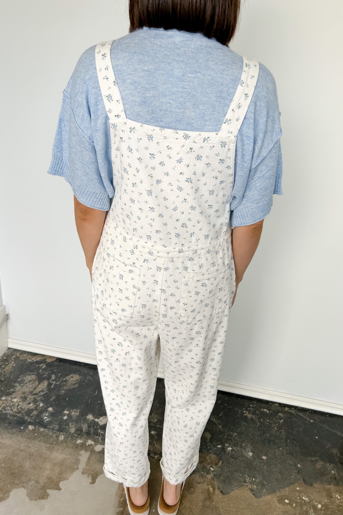 Person wearing white floral overalls and a blue shirt against a plain background
