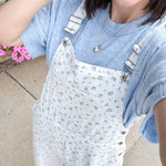 Person wearing a light blue sweater and white floral overalls standing outdoors with flowers in the background.