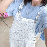 Person wearing a light blue sweater and white floral overalls standing outdoors with flowers in the background.