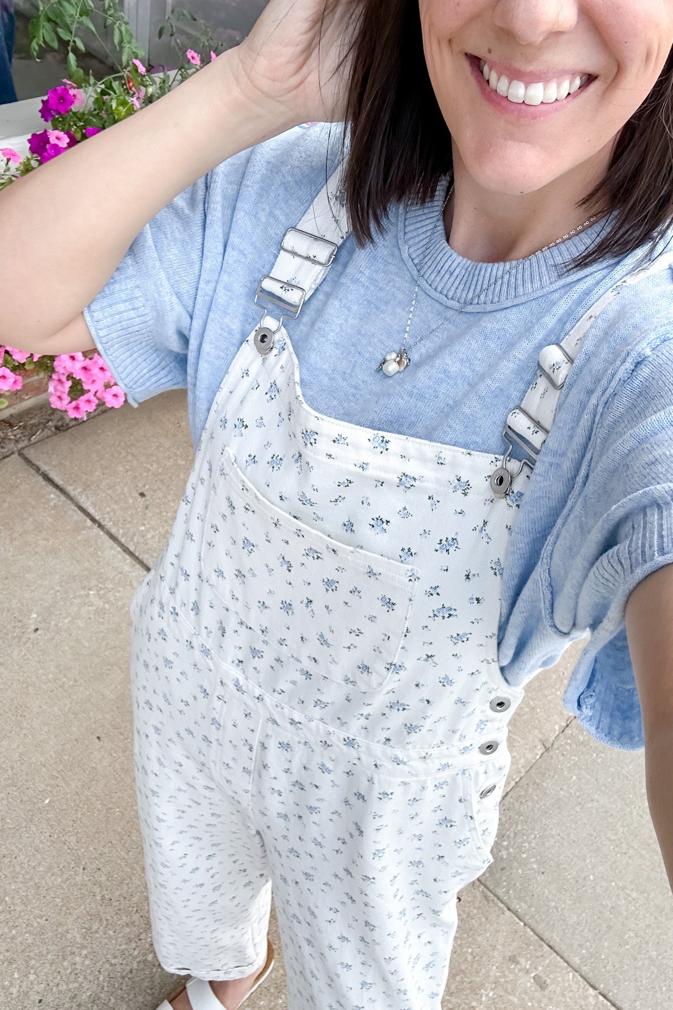 Person wearing a light blue sweater and white floral overalls standing outdoors with flowers in the background.