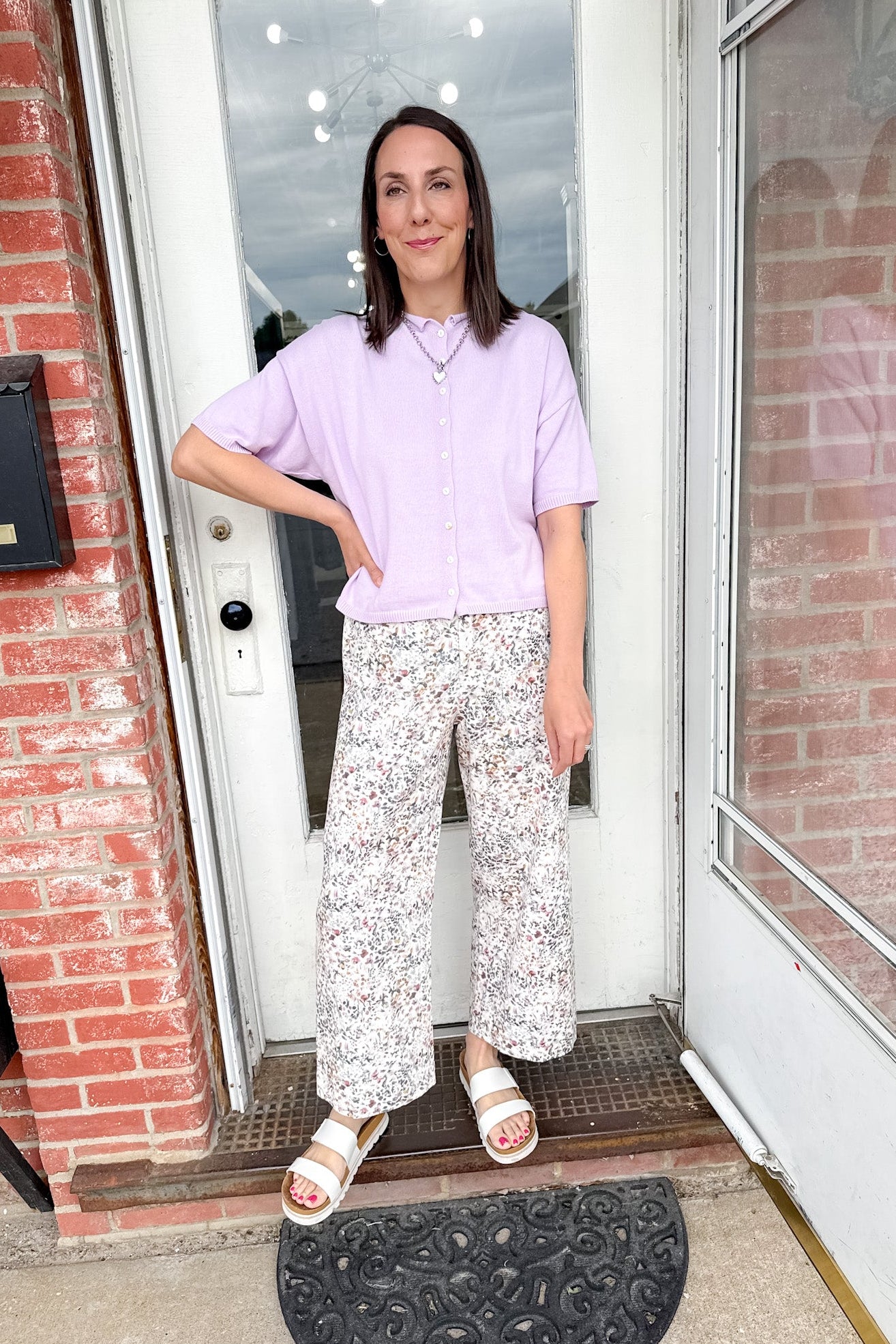 Woman standing in front of a door wearing a light purple shirt and white floral pants.