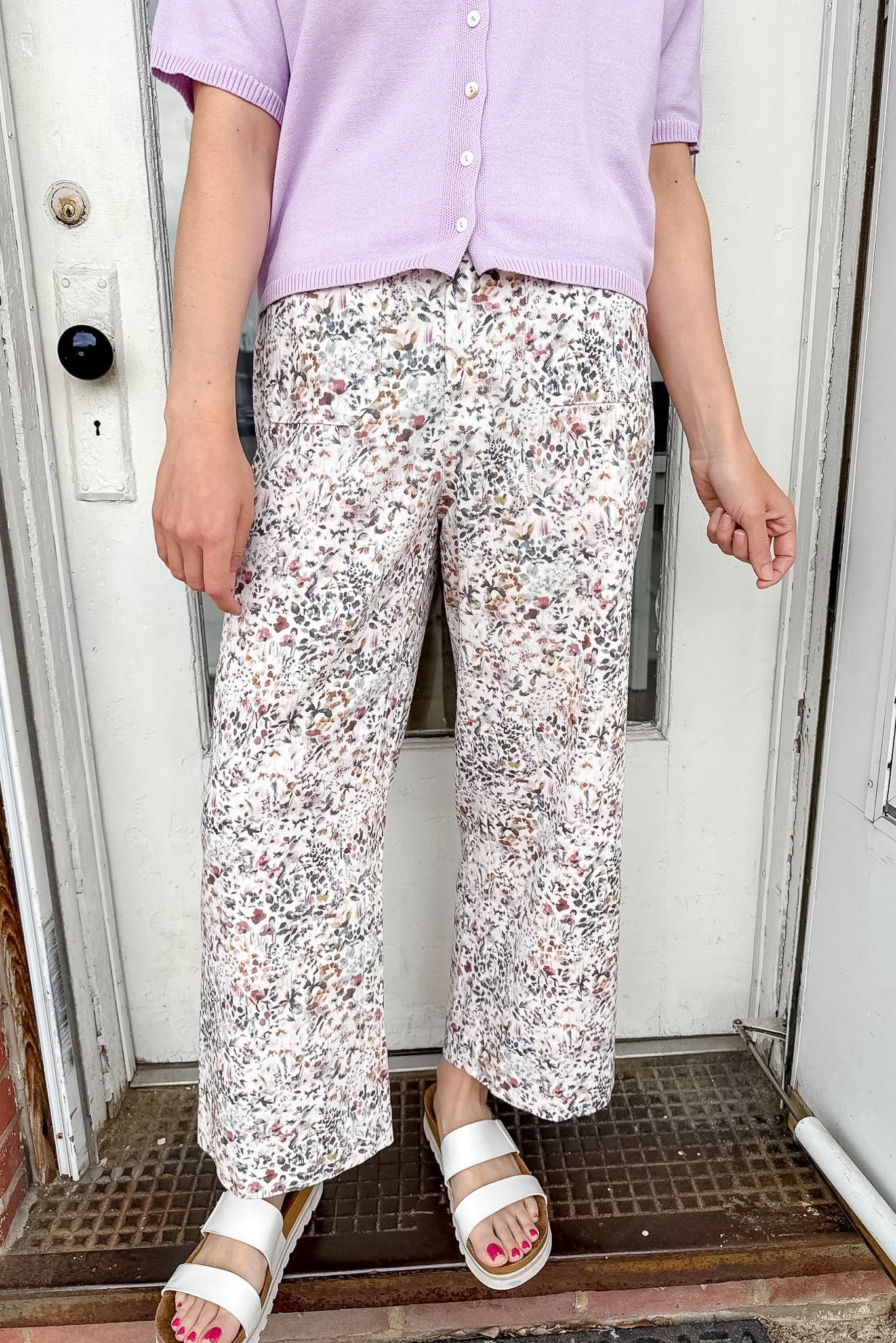 Person wearing a light purple shirt and floral pants standing in front of a door.