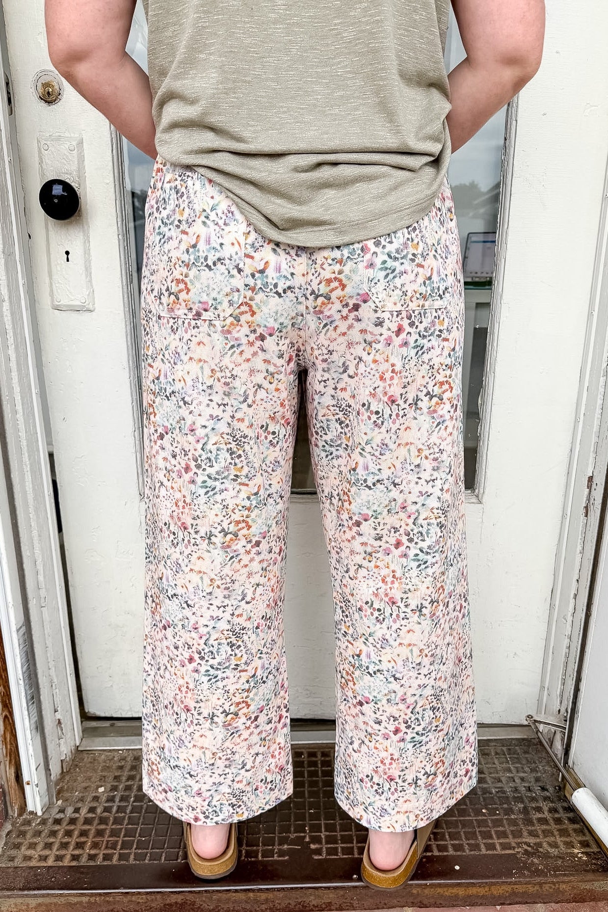 Person wearing floral pants and a green top standing in front of a door.