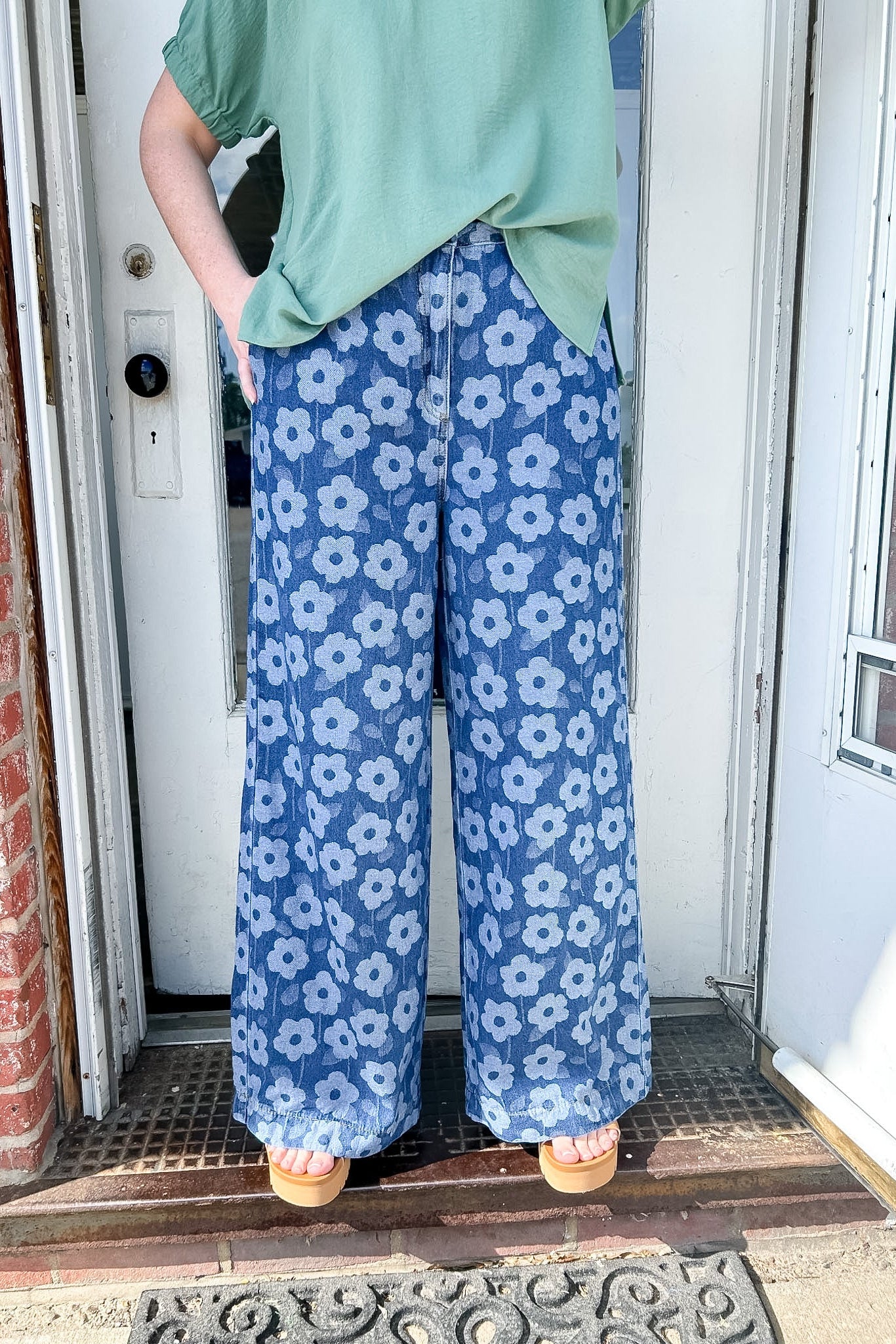 Person wearing blue floral pants and a green top standing in front of a door.