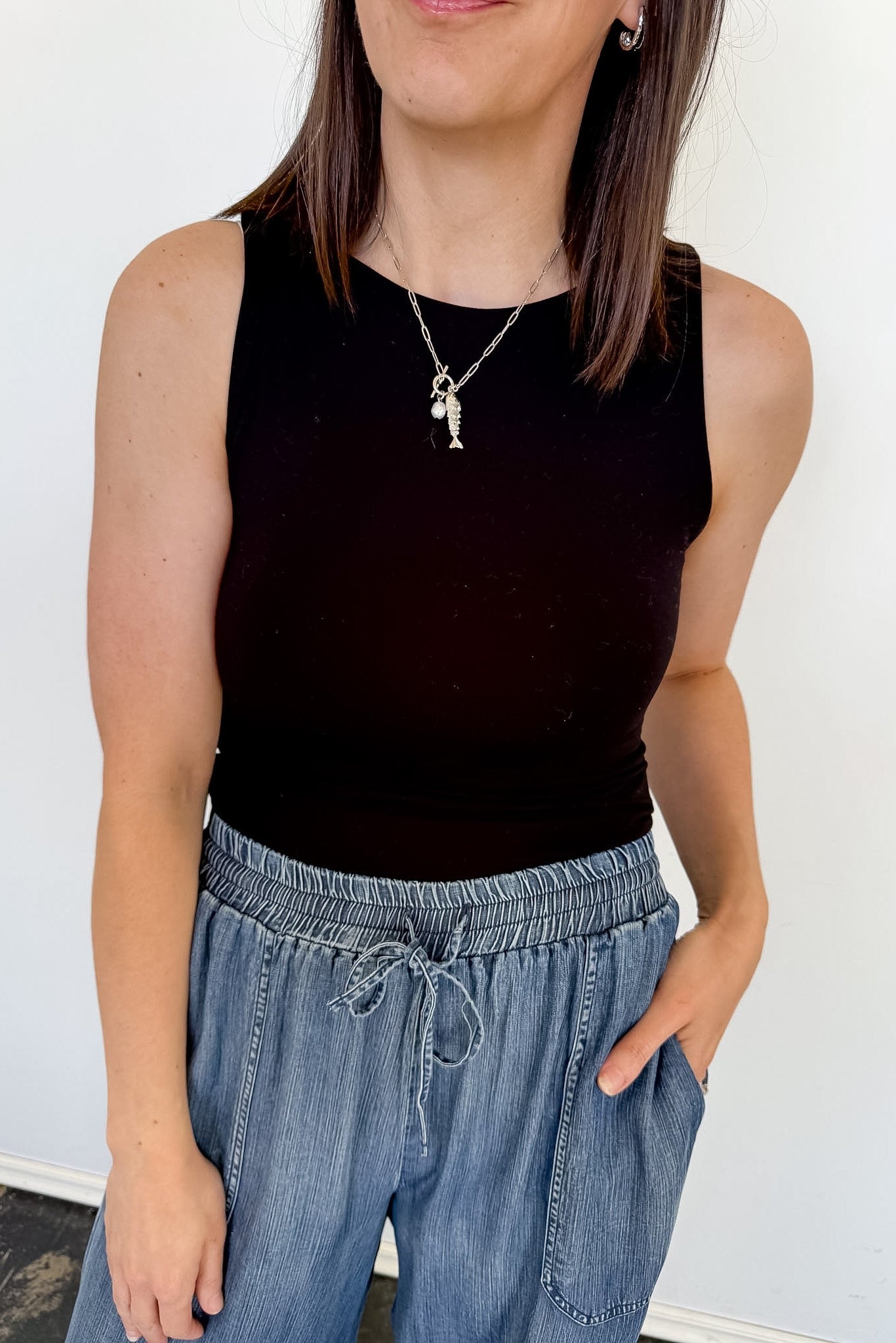 Person wearing a black sleeveless top and blue jeans against a white wall
