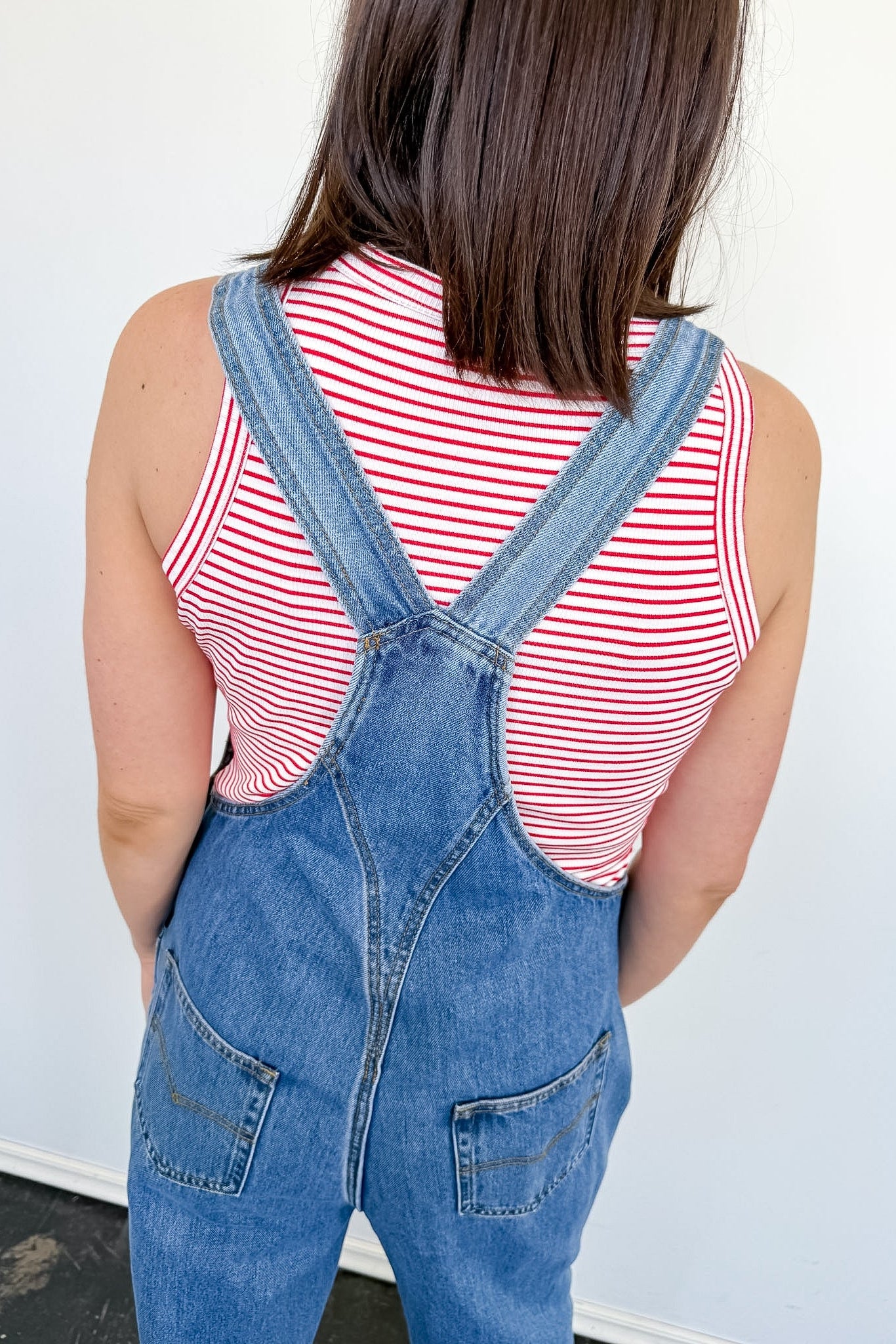 A woman standing in front of a white wall with her back to the camera. She is wearing a red and white striped tank with overalls.