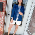 Woman standing in front of a door wearing a blue jacket, striped shirt, and white shorts.