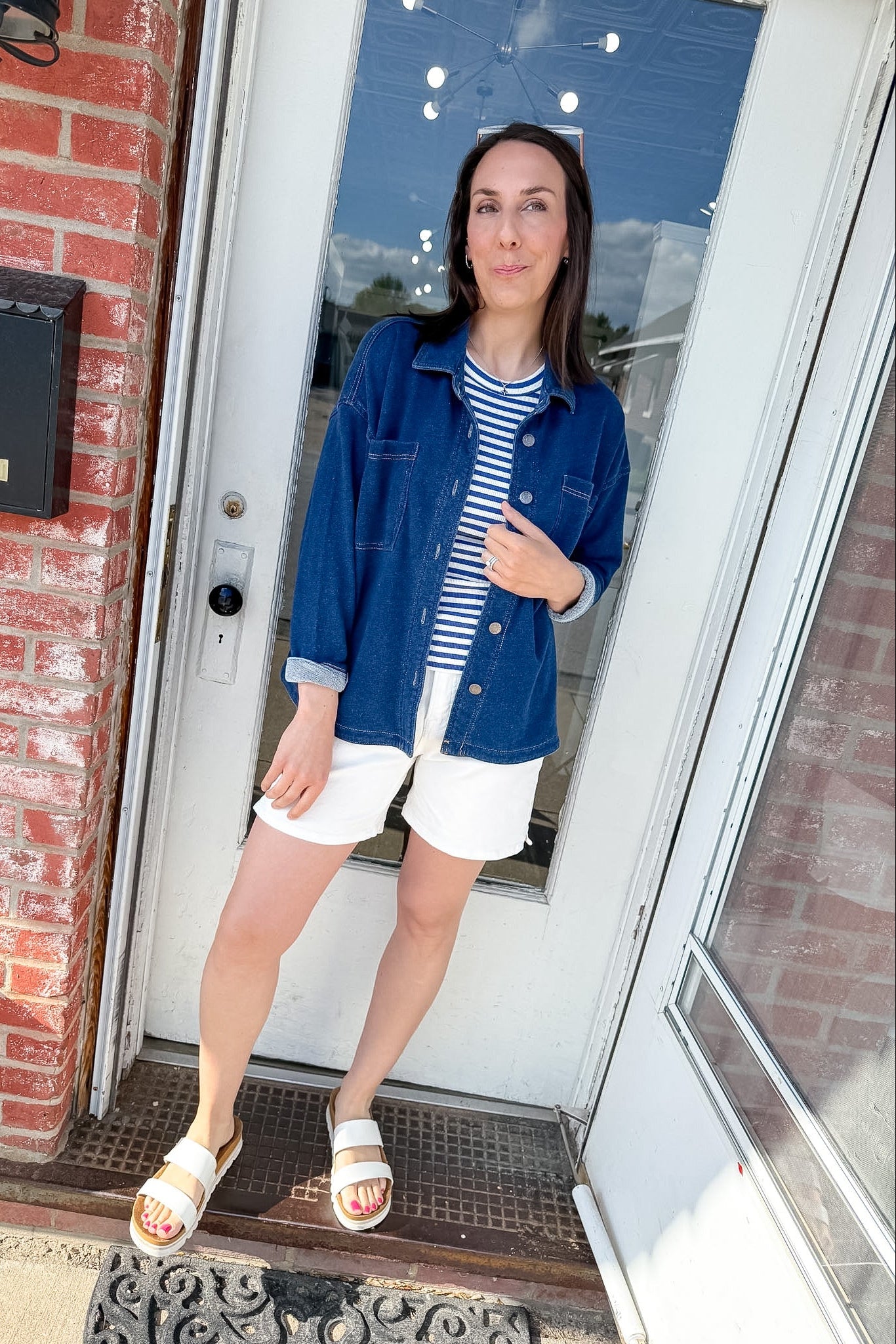 Woman standing in front of a door wearing a blue jacket, striped shirt, and white shorts.