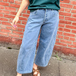 Person wearing a green shirt and blue jeans standing against a red brick wall.