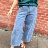 Person wearing a green shirt and blue jeans standing against a red brick wall.