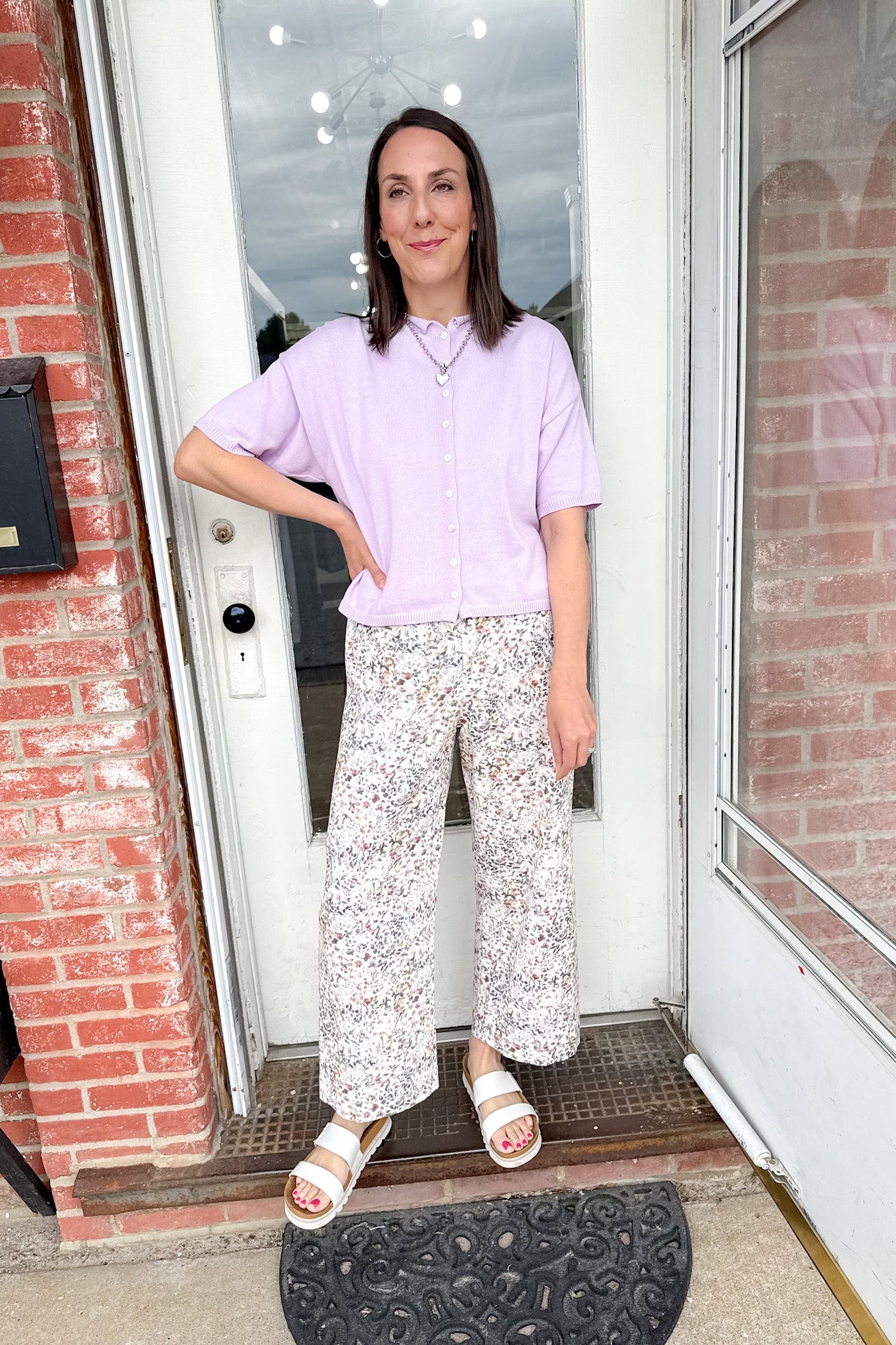 Woman standing in front of a door wearing a light purple shirt and white floral pants.