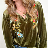 Woman wearing an olive green velvet blouse with floral embroidery on a white background