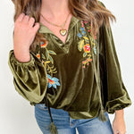 Woman wearing an olive green velvet top with floral embroidery and blue jeans against a white wall.