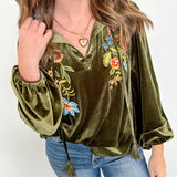 Woman wearing an olive green velvet top with floral embroidery and blue jeans against a white wall.