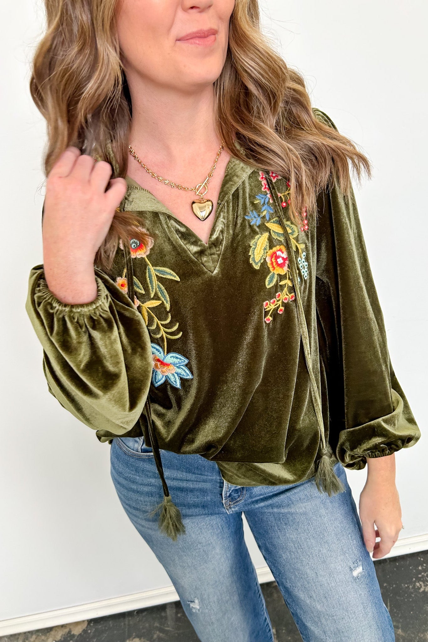 Woman wearing an olive green velvet top with floral embroidery and blue jeans against a white wall.