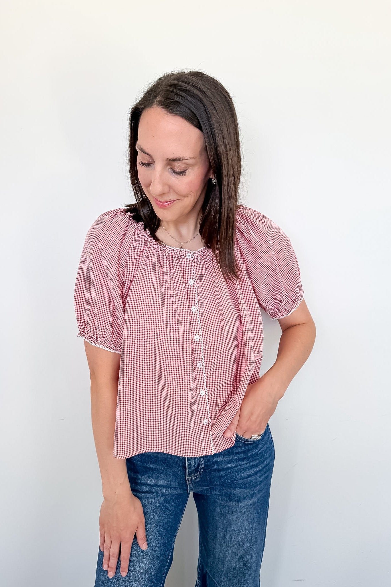 A woman in a red and white gingham top with puff sleeves standing in front of white wall. 