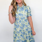 Woman wearing a blue and green floral dress against a white wall.