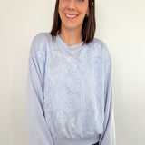 Woman wearing a light blue tie-dye sweatshirt and jeans against a white wall.