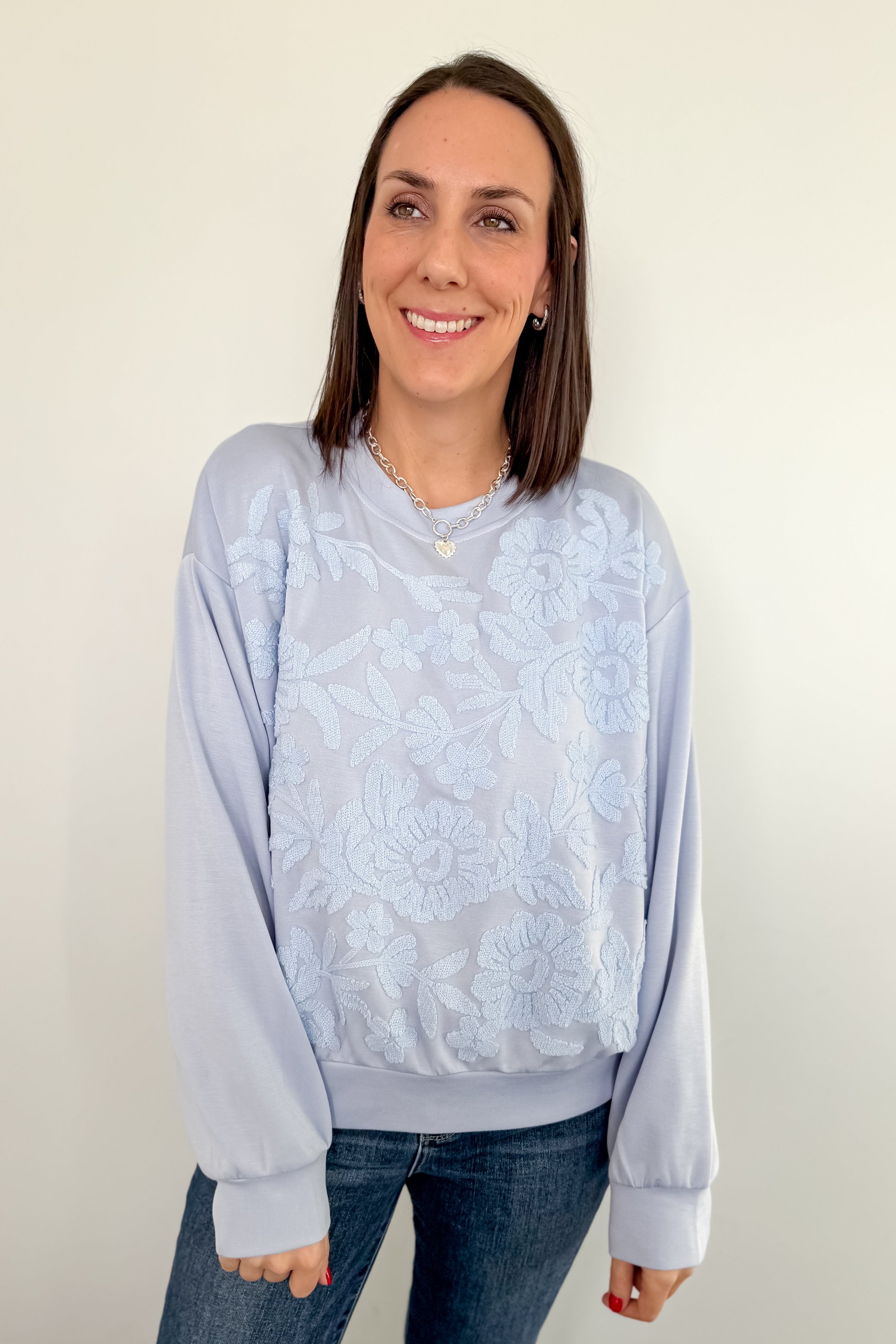Woman wearing a light blue tie-dye sweatshirt and jeans against a white wall.