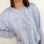 Person wearing a light gray sweatshirt with floral lace overlay on a plain background